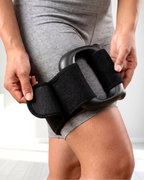 A person wearing gray athletic shorts is shown from the waist down, holding a black thigh support brace around their upper thigh in a bright indoor room.